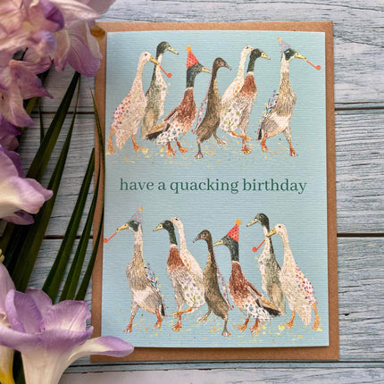 Greeting card with ducks illustration and 'have a quacking birthday' text on a wooden surface with flowers.