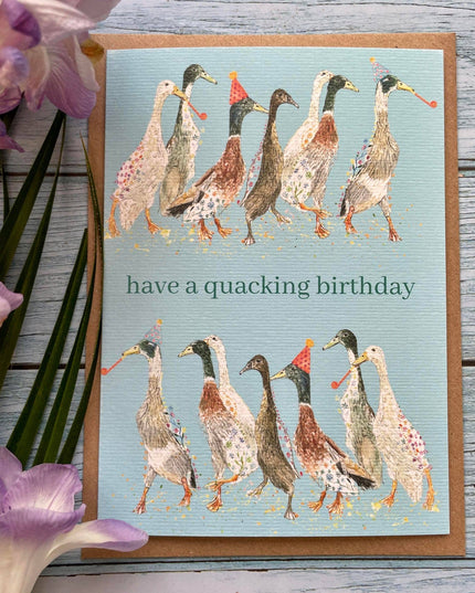 Greeting card with ducks illustration and 'have a quacking birthday' text on a wooden surface with flowers.