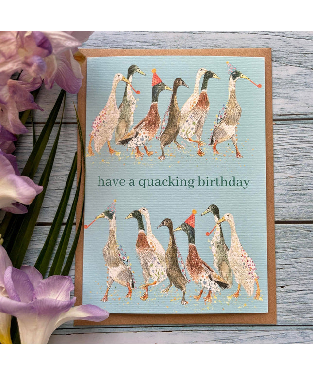 Greeting card with ducks illustration and 'have a quacking birthday' text on a wooden surface with flowers.
