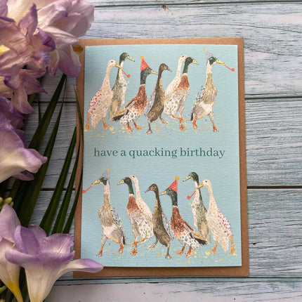 Greeting card with ducks illustration and 'have a quacking birthday' text on a wooden surface with flowers.