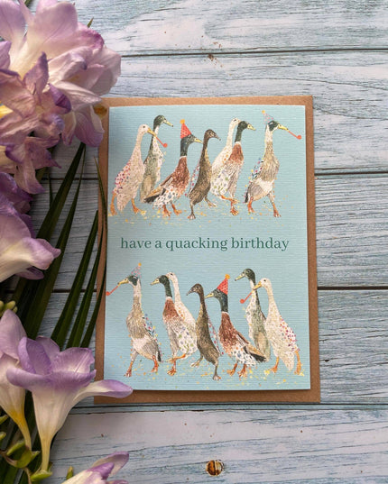 Greeting card with ducks illustration and 'have a quacking birthday' text on a wooden surface with flowers.