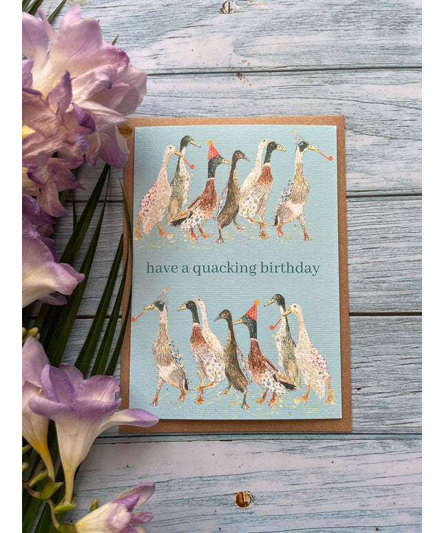 Greeting card with ducks illustration and 'have a quacking birthday' text on a wooden surface with flowers.