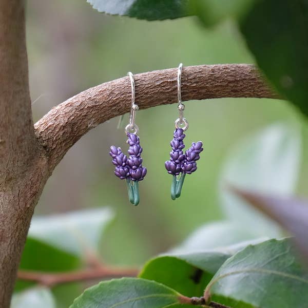 A pair of dangling earrings shaped like small purple flower clusters with green stems, hanging from a tree branch outdoors with blurred green leaves in the background.