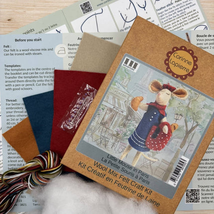 A felt craft kit package labeled 'Corinne Laquenne' and 'Little Mouse in Paris' lies on top of several printed instruction sheets. The package shows a felted mouse figure in blue and red clothing, with wool roving and folded felt pieces visible around it.