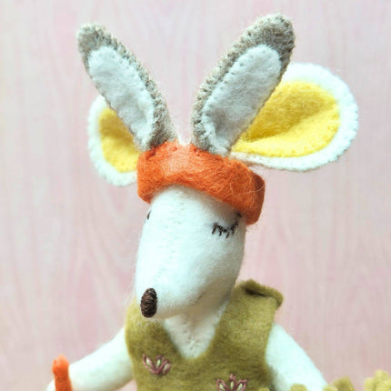 A close-up of a handmade felt animal toy, likely a rabbit or kangaroo, with long white and gray ears, yellow inner ears, an orange headband, and a green vest, set against a soft pink background.