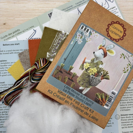 A wool felt craft kit package for 'Little Mouse is a Bunny' by Corinne Laperre, displayed on top of other craft materials including yarn, paper templates, and wool on a light wood surface.