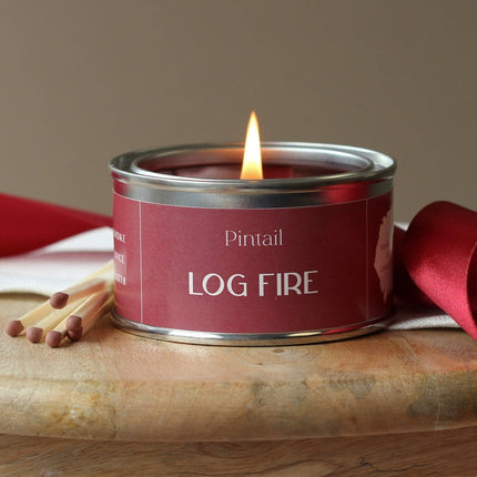 Candle in a red tin labeled 'Log Fire' by Pintail, with matches on a wooden surface.