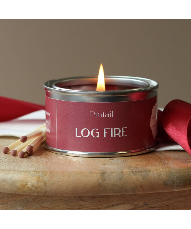 Candle in a red tin labeled 'Log Fire' by Pintail, with matches on a wooden surface.