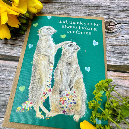 Greeting card with meerkats and flowers on a wooden surface with yellow flowers.