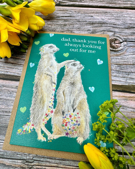 Greeting card with meerkats and flowers on a wooden surface with yellow flowers.
