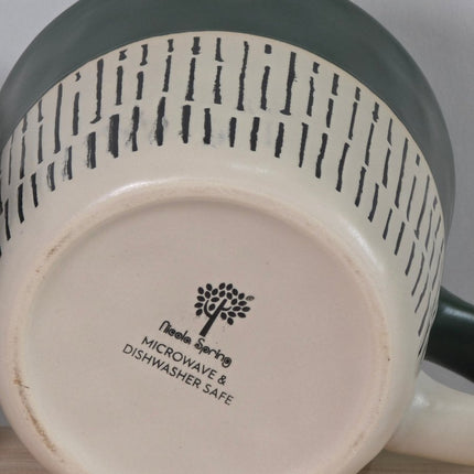 Close-up of the bottom of a ceramic mug or pitcher, showing a cream-colored base with black printed text that reads Microwave & Dishwasher Safe' alongside a tree logo. 