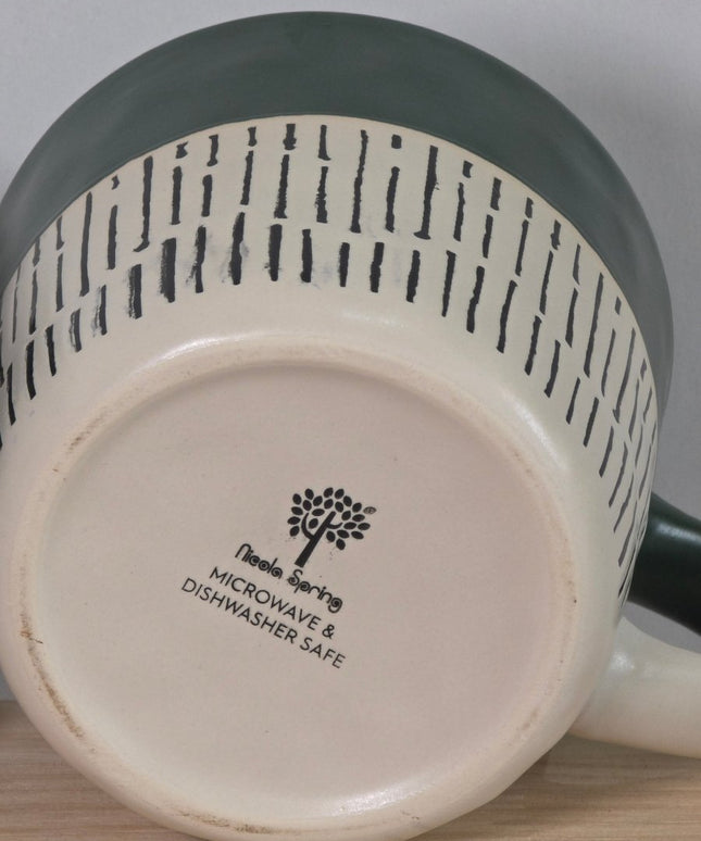 Close-up of the bottom of a ceramic mug or pitcher, showing a cream-colored base with black printed text that reads Microwave & Dishwasher Safe' alongside a tree logo. 