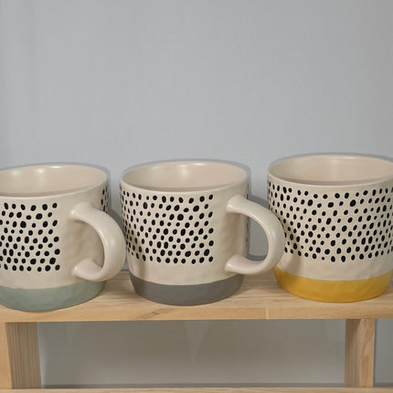Three ceramic mugs with a black dotted pattern and different colored bases, displayed on a light wooden stand.