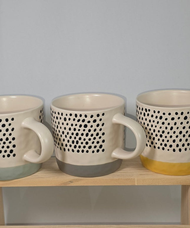 Three ceramic mugs with a black dotted pattern and different colored bases, displayed on a light wooden stand.