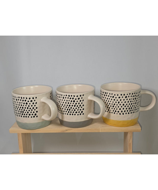 Three ceramic mugs with a black dotted pattern and different colored bases, displayed on a light wooden stand.
