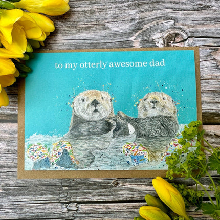 Greeting card with two otters and text 'to my otterly awesome dad' on a wooden surface with yellow flowers.
