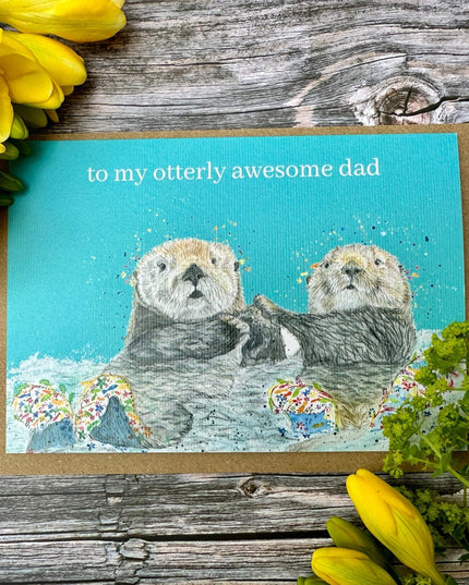 Greeting card with two otters and text 'to my otterly awesome dad' on a wooden surface with yellow flowers.