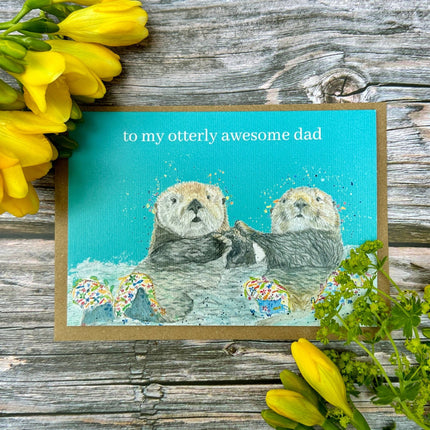 Card with two otters and text 'to my otterly awesome dad' on a wooden background with yellow flowers.