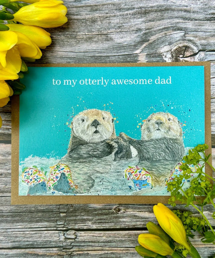 Card with two otters and text 'to my otterly awesome dad' on a wooden background with yellow flowers.