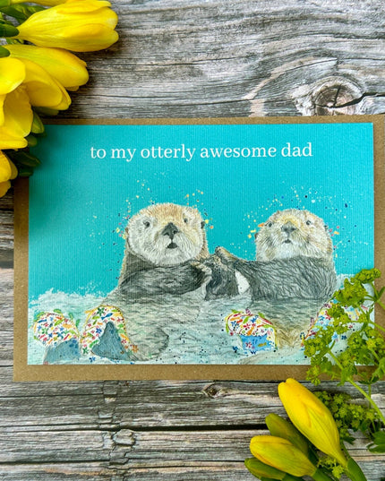 Card with two otters and text 'to my otterly awesome dad' on a wooden background with yellow flowers.