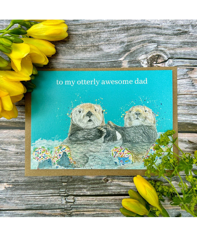 Card with two otters and text 'to my otterly awesome dad' on a wooden background with yellow flowers.