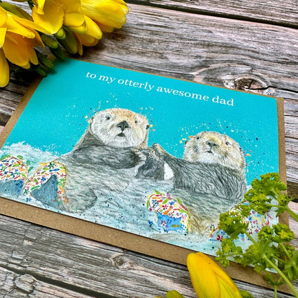 Card with two otters and text 'to my otterly awesome dad' on a wooden surface with yellow flowers.