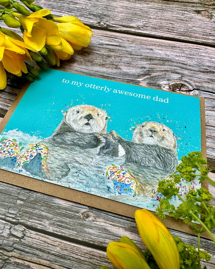 Card with two otters and text 'to my otterly awesome dad' on a wooden surface with yellow flowers.