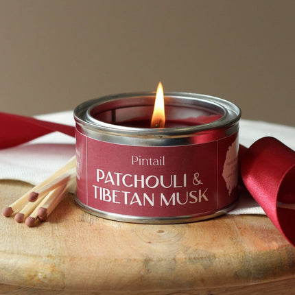 Candle in a red tin labeled 'Pintail Patchouli & Tibetan Musk' with matches and a red ribbon on a wooden surface.