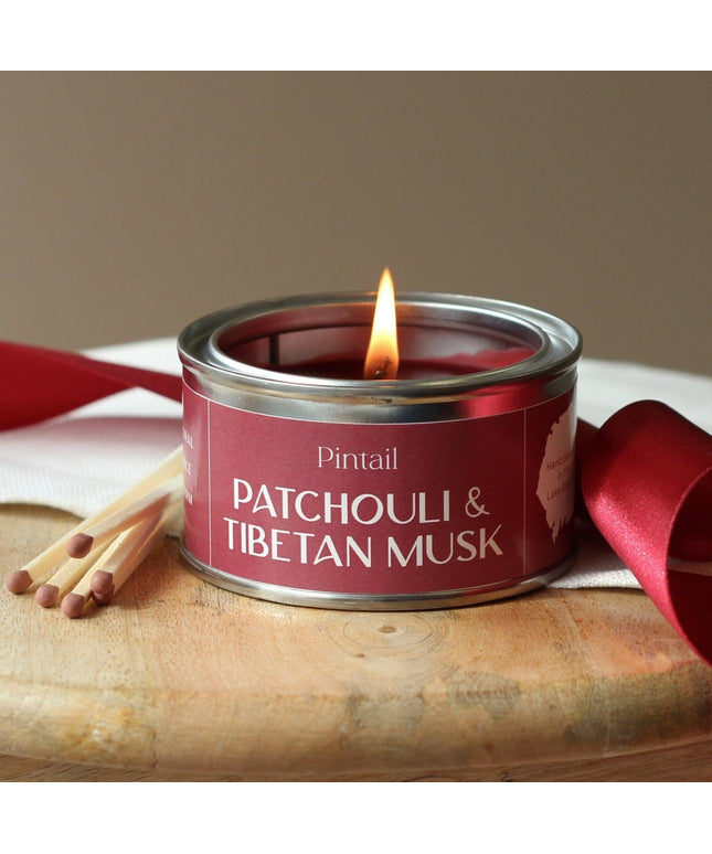 Candle in a red tin labeled 'Pintail Patchouli & Tibetan Musk' with matches and a red ribbon on a wooden surface.
