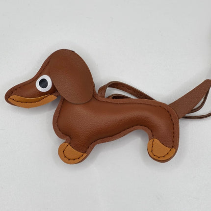A brown faux-leather dachshund-shaped keychain or bag charm with tan accents on the muzzle, paws, and tail tip, and a black-and-white plastic eye, shown on a plain light background.