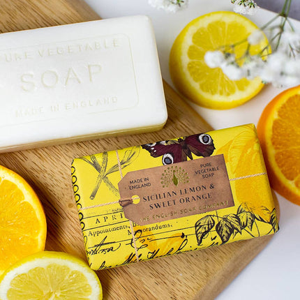 Two bars of soap, one white and one yellow with lemon and orange design, on a wooden board with sliced lemons and oranges.