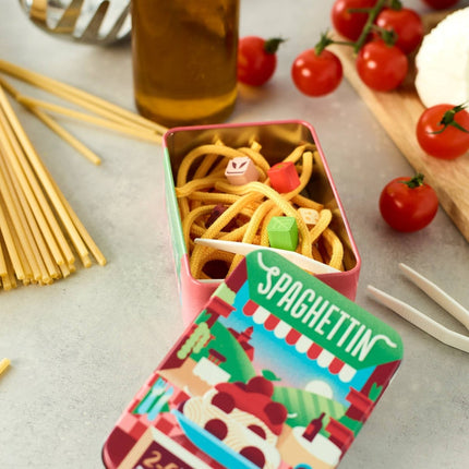 A colorful metal tin of spaghetti pasta with a matching lid placed in front. The tin is styled on a light gray surface with whole wheat spaghetti sticks, a bottle of olive oil, cherry tomatoes, and cheese in a kitchen setting.