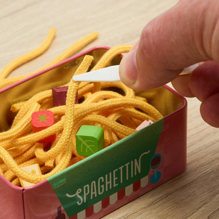 A close-up of a hand using a white plastic pick to remove yellow spaghetti-shaped cords from a pink and green tin labeled 'SPAGHETTIN', with small colorful cube-shaped pieces visible inside.