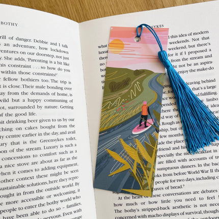 Book with an illustrated bookmark featuring a person on a surfboard, surrounded by nature, on an open book.