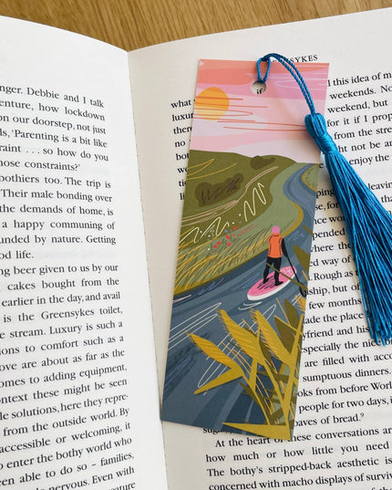 Book with an illustrated bookmark featuring a person on a surfboard, surrounded by nature, on an open book.