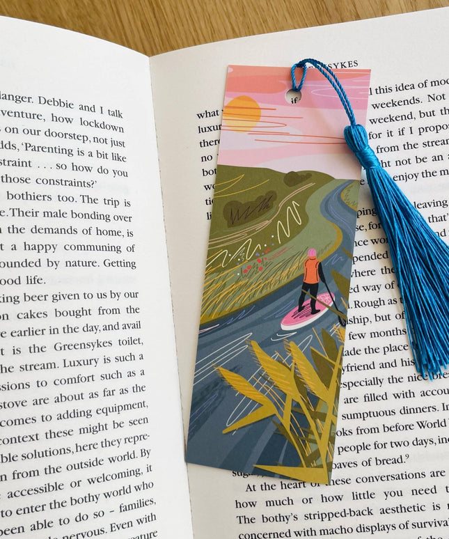 Book with an illustrated bookmark featuring a person on a surfboard, surrounded by nature, on an open book.