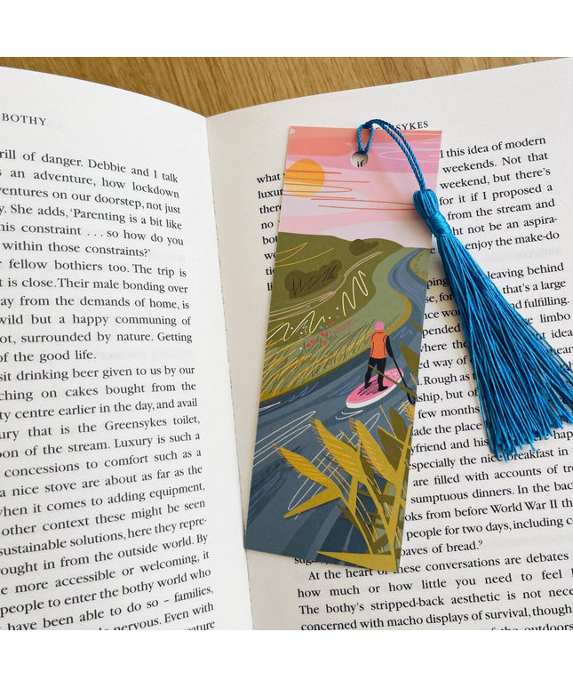 Book with an illustrated bookmark featuring a person on a surfboard, surrounded by nature, on an open book.